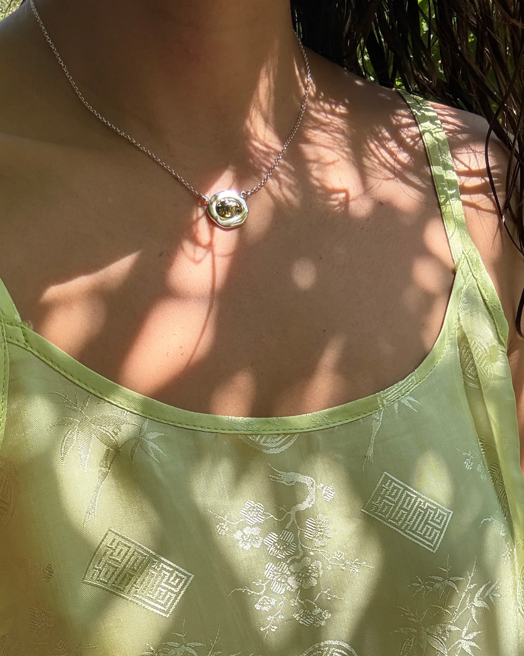 A close-up lifestyle shot of a person wearing a delicate silver necklace with a round stone pendant, paired with a light sage green camisole top featuring white embroidered patterns and a subtle bamboo motif.