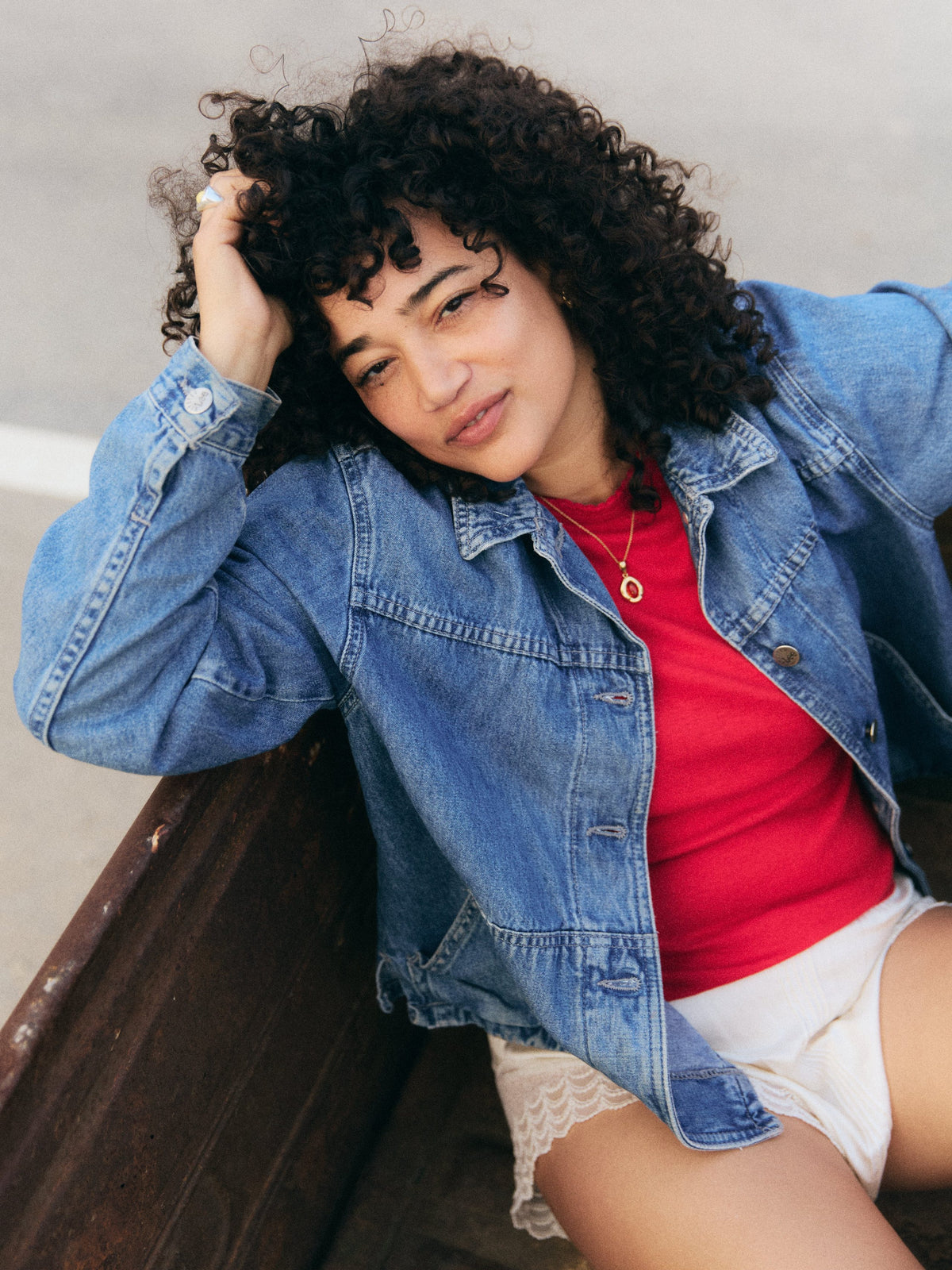 Woman in a denim jacket and red top leaning on a bench, wearing a gold Baltic amber pendant necklace