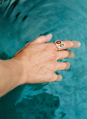 Hand trailing through turquoise water, wearing the Molten Trio Ring with multicolor Baltic amber