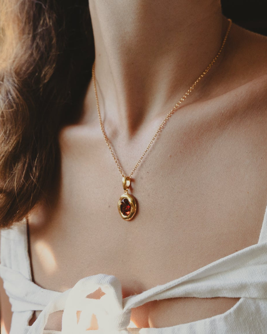 A close-up lifestyle shot of a woman wearing a delicate gold chain necklace with a small, dark teardrop-shaped pendant, paired with a white sleeveless top.