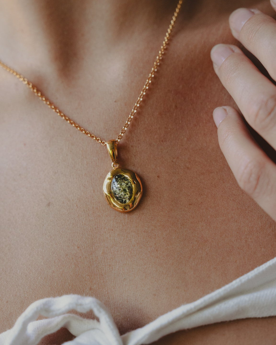 A gold chain necklace with a small oval pendant featuring a green and gold pattern, worn on a model's neck above a white tie-front top.
